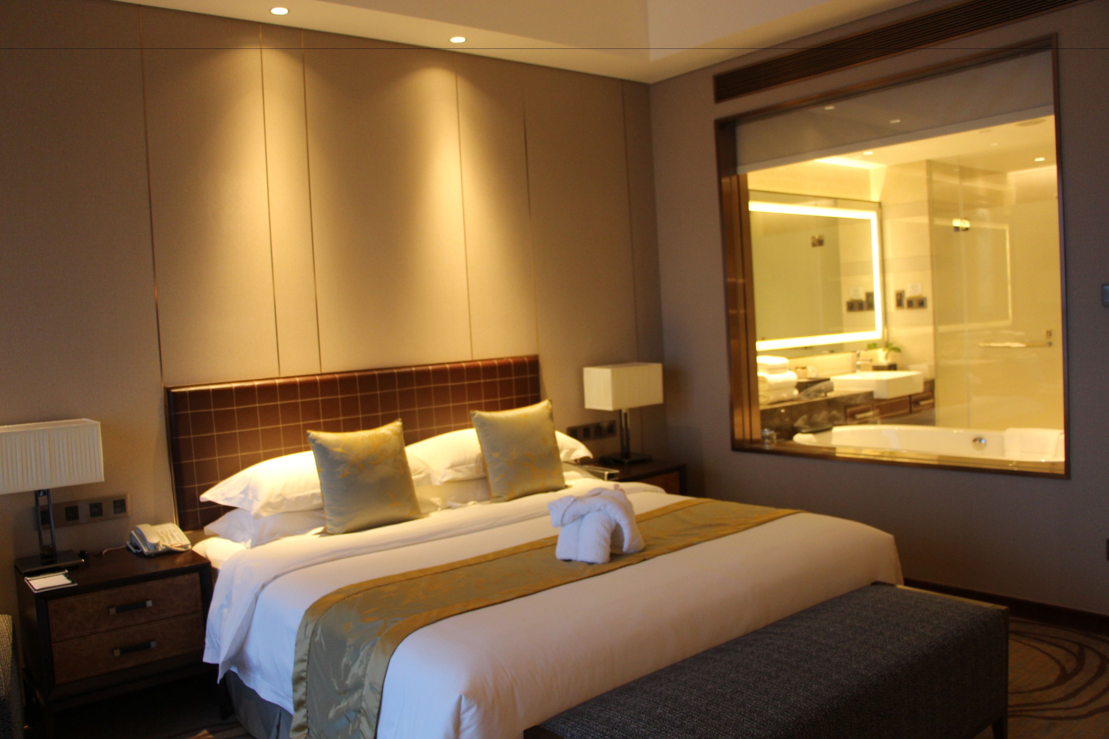 Habitación de hotel con una cama grande, una mesita de noche con lámpara, un espejo y un baño visible a través de la pared de vidrio.