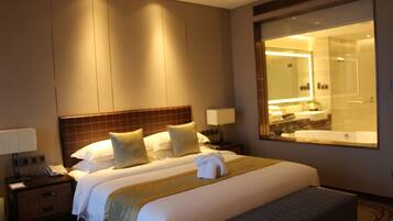 Habitación de hotel con una cama grande, una mesita de noche con lámpara, un espejo y un baño visible a través de la pared de vidrio.