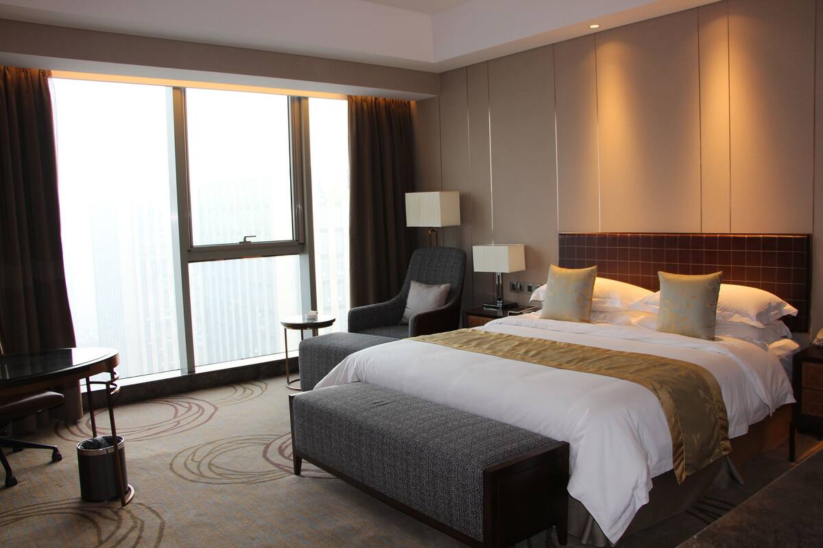 Habitación de hotel con una cama grande, un escritorio, una silla y vistas a la ciudad a través de amplios ventanales.