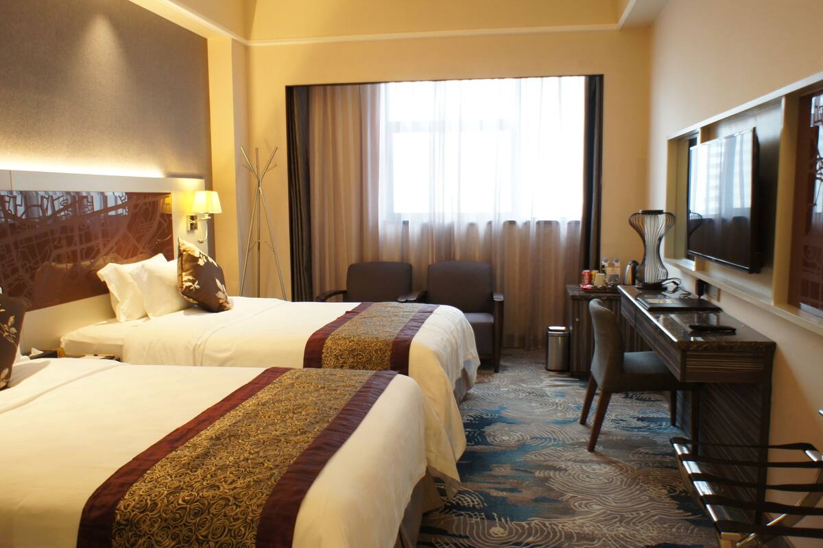 A hotel room with two beds, a desk, a chair, a TV, and a window with curtains.