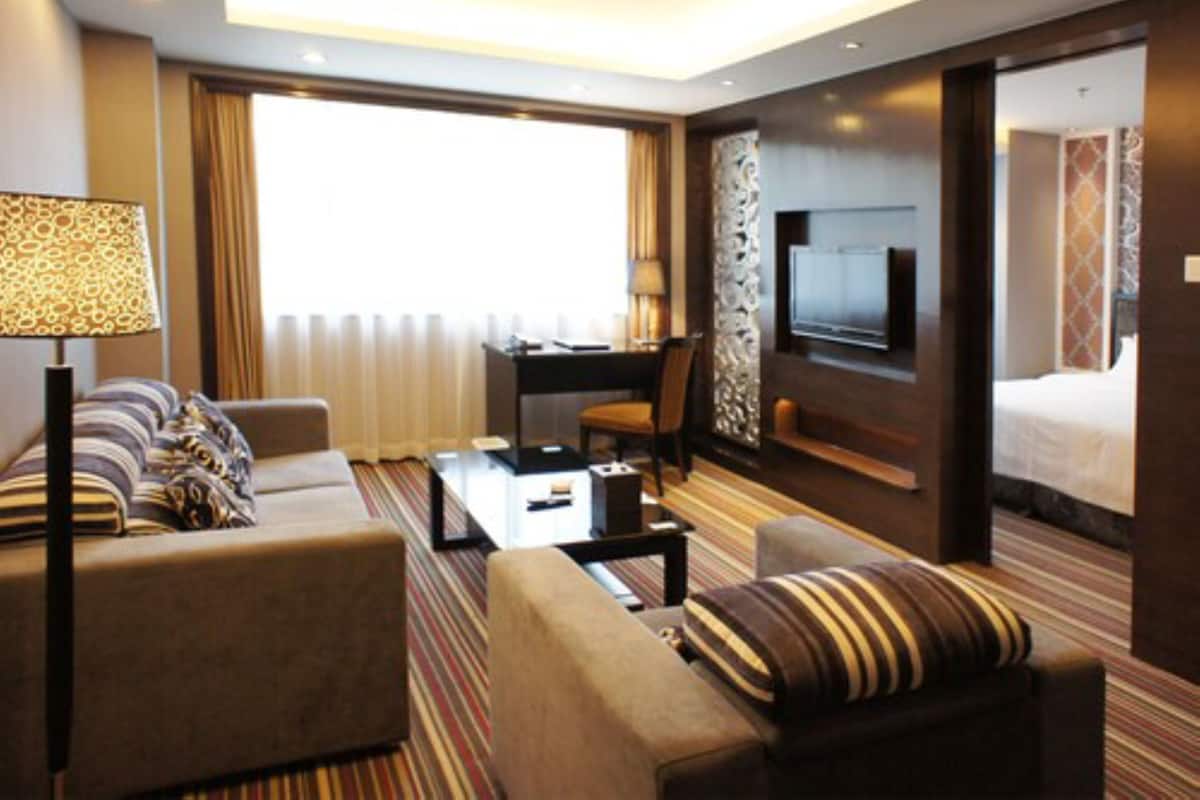A hotel room with a sofa set, a coffee table, a desk with a chair, a TV mounted on the wall, and a bed.