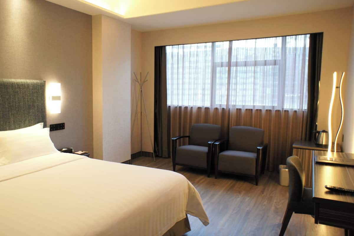 A hotel room with a large bed, a desk with a chair, a lamp, and a window with curtains.