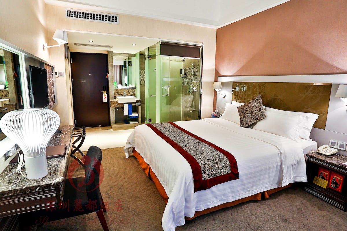 A hotel room with a bed, a desk with a lamp, a chair, a bedside table with a phone, and a shower area.