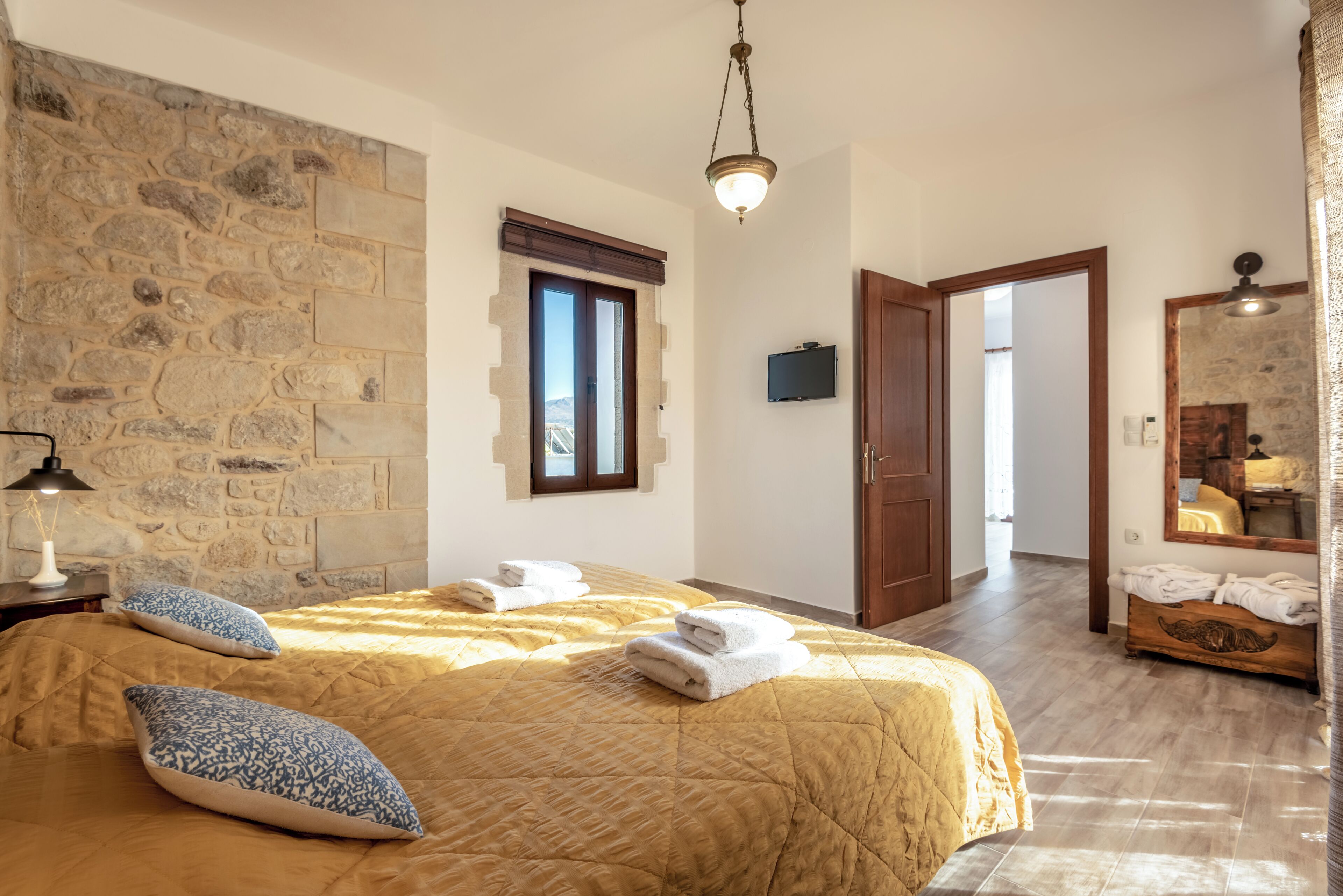 Un dormitorio con pared de piedra, una cama con colcha, dos almohadas, una lámpara de noche, un televisor y un aparador de madera.