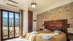 A bedroom with a large bed, a stone wall, a wooden headboard, and a balcony with a view of the mountains. - Villa Aerino (Kissamos)
