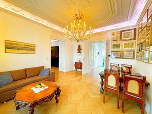 A room with a chandelier, a wooden dining table with chairs, a sofa, and a framed picture on the wall.