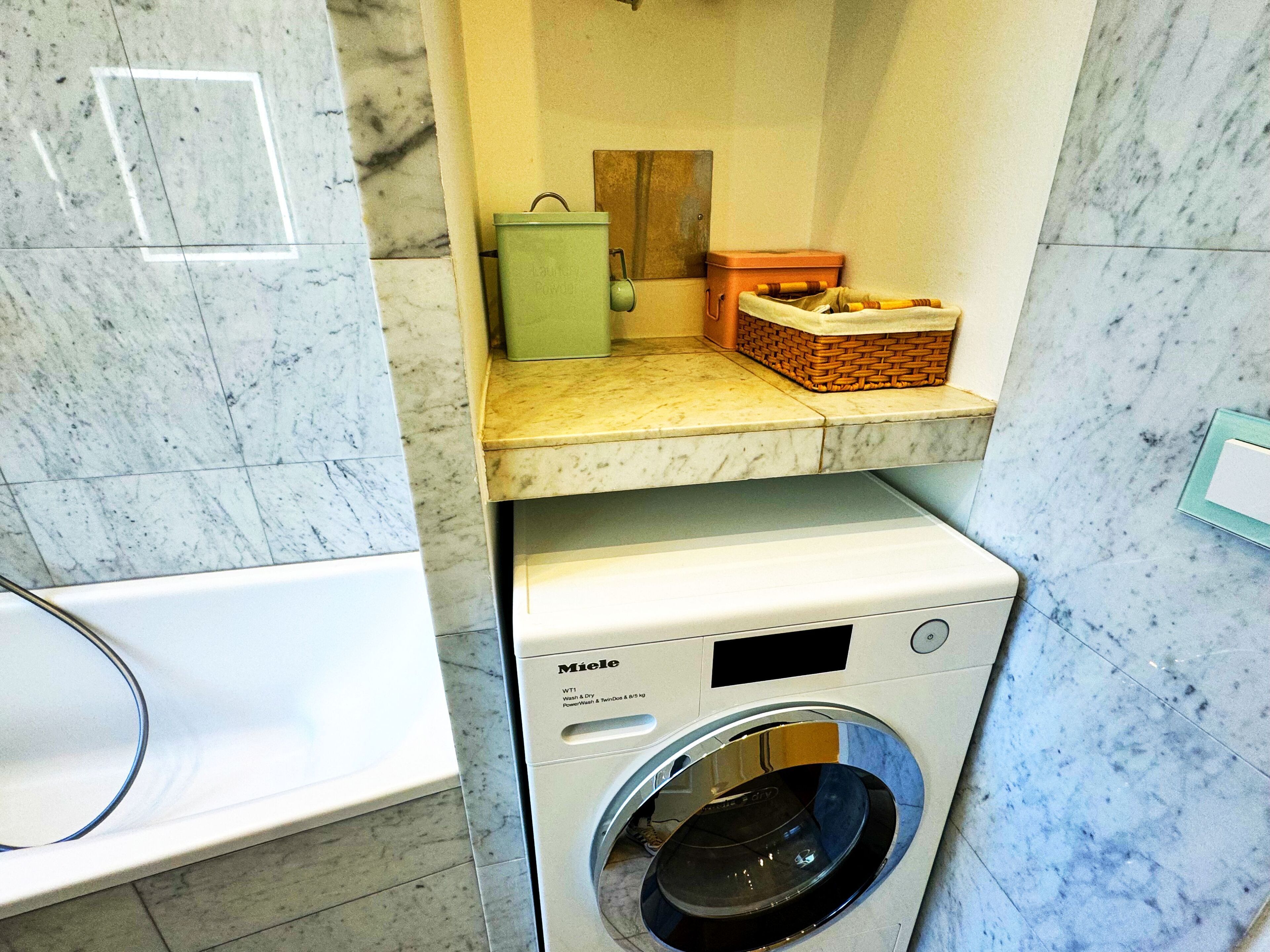 Un lavadero compacto y moderno con lavadora Miele de carga frontal, una caja verde pequeña y una cesta de mimbre sobre una encimera de mármol.