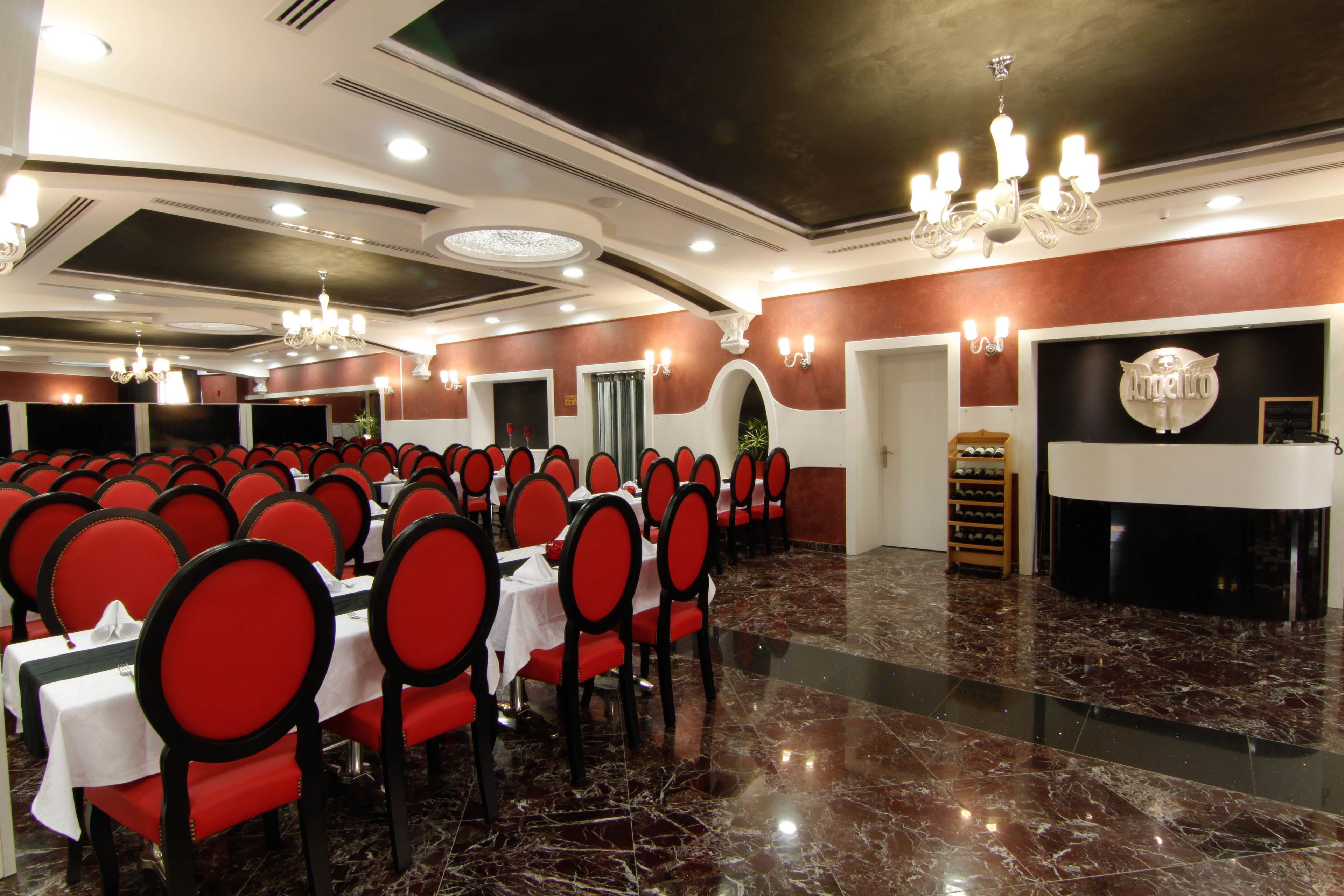 Un salón de banquetes con filas de mesas y sillas, una araña y una zona de recepción con mostrador y cartel.