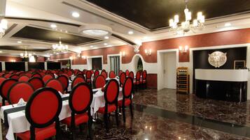 Un salón de banquetes con filas de mesas y sillas, una araña y una zona de recepción con mostrador y cartel.