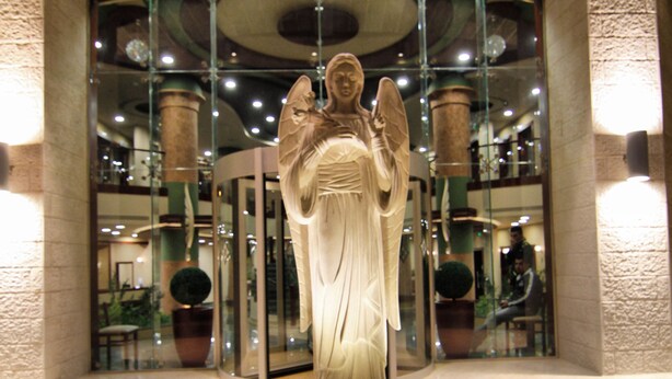 A statue of an angel on a circular pedestal in a lobby with a glass elevator and potted plants. - Saint Gabriel Hotel (Bethlehem)