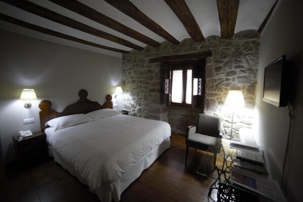 Un dormitorio con una cama grande, vigas de madera, paredes de piedra, un escritorio y dos lámparas.