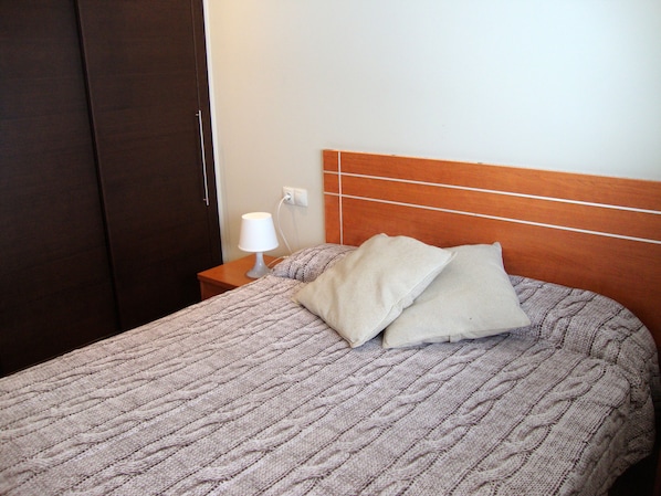 Ein ordentlich bezogenes Bett mit Strickdecke und zwei Kissen, einer Nachttischlampe und einem hölzernen Kopfteil.