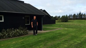 A black building with a number '8' on the side, a grassy lawn, and a hedge with white flowers. - Blokhus Golfcenter (Pandrup)