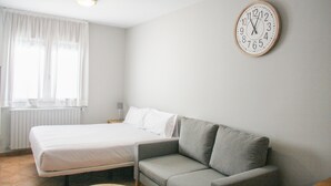 A room with a bed, a grey sofa, a wall clock, and a window with curtains. - Aptos Colells (Bordes d'Envalira)