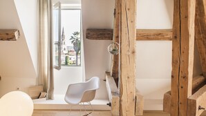 Una habitación con vigas de madera a la vista, una silla blanca, una mesita y vista a un edificio con una torre del reloj a través de una ventana.