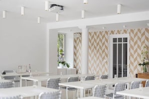 A modern dining area with white tables and chairs, a patterned wall, and a large window with plants. - La Villa Guy (Béziers)