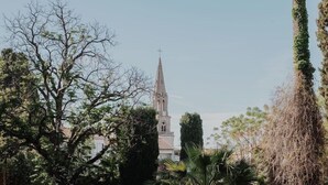 A well-maintained garden with a variety of trees and shrubs, a clear pathway leading to a building with a spire, and a distant view of a church. - La Villa Guy (Béziers)