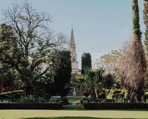 A well-maintained garden with a variety of trees and shrubs, a clear pathway leading to a building with a spire, and a distant view of a church. - La Villa Guy (Béziers)