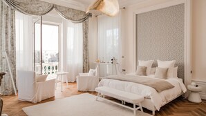Un dormitorio con una cama grande, un banco, dos sillas, una mesita con una lámpara y vistas al exterior.