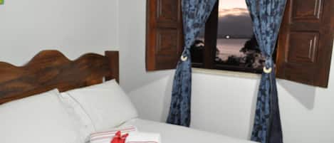 Uma cama com lençóis brancos, cabeceira de madeira e janela com cortinas azuis.