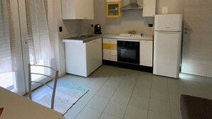 A compact kitchen with white cabinets, a stove, a sink, and a refrigerator. There is a window with curtains and a chair in the foreground. - Le Sorbole (Matera)