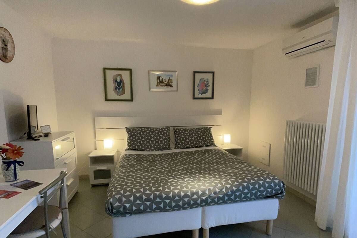 Une chambre à coucher comprenant un lit, des tables de chevet, un bureau et des chaises. Les murs sont ornés de tableaux encadrés et la pièce est équipée d’un climatiseur.