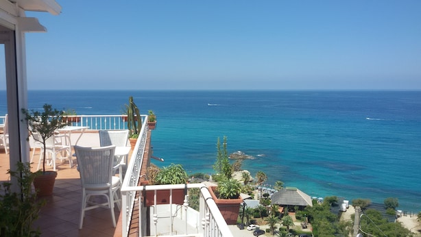 Ein Balkon mit weißen Korbstühlen und Topfpflanzen mit Blick auf einen Strand und klares blaues Wasser sowie Boote.