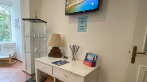 Ein Zimmer mit weißem Schreibtisch, einem an der Wand befestigten Fernseher, einem Schild, das eine Lounge Bar kennzeichnet, einer Glasvitrine, einer Lampe und einer Topfpflanze.