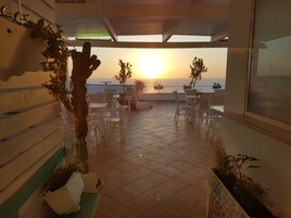 Um terraço com mobiliário de vime branco, plantas em vasos e vista para o pôr do sol sobre o mar.