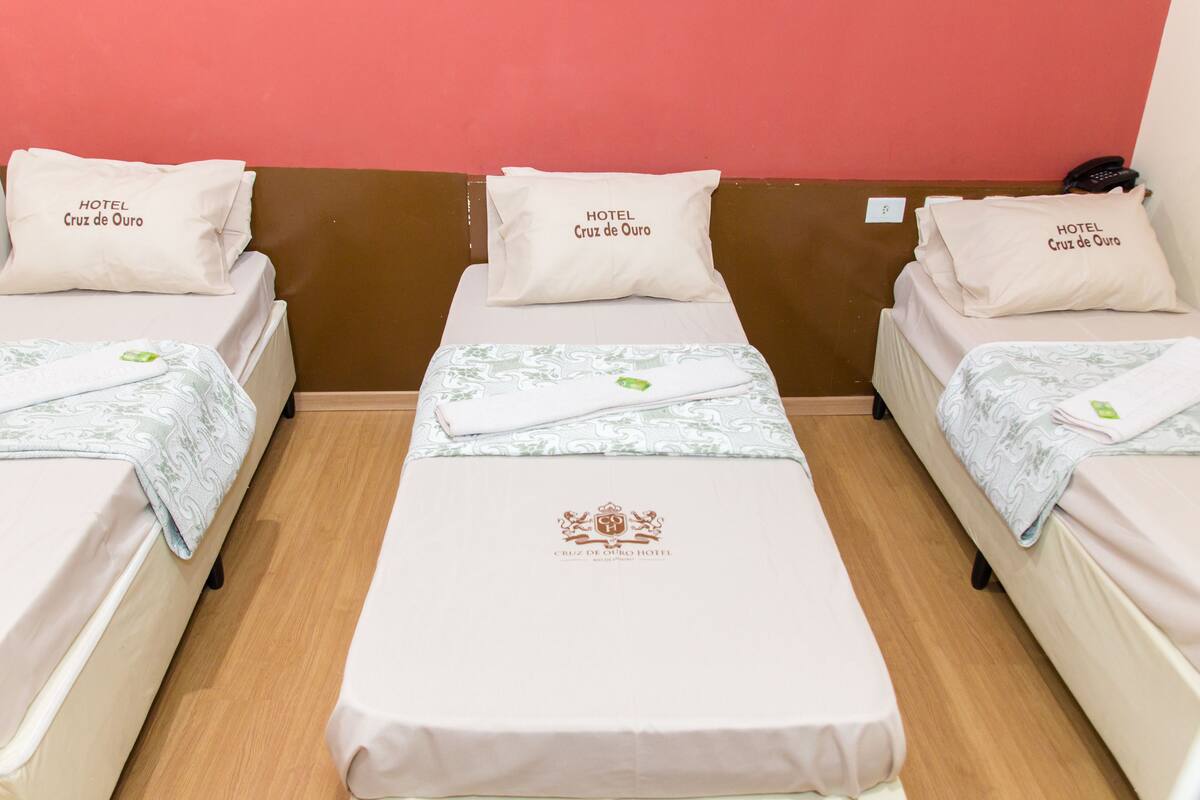 A hotel room with three single beds, each with a pillow labeled "HOTEL Cruz de Ouro". The beds have a floral-patterned bedspread and a central bed has a logo with the hotel name.
