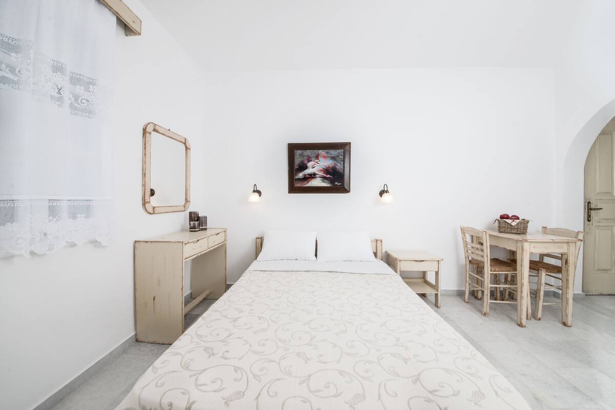 A bedroom with a bed, a desk, a chair, a small table, a mirror, and a framed picture on the wall.