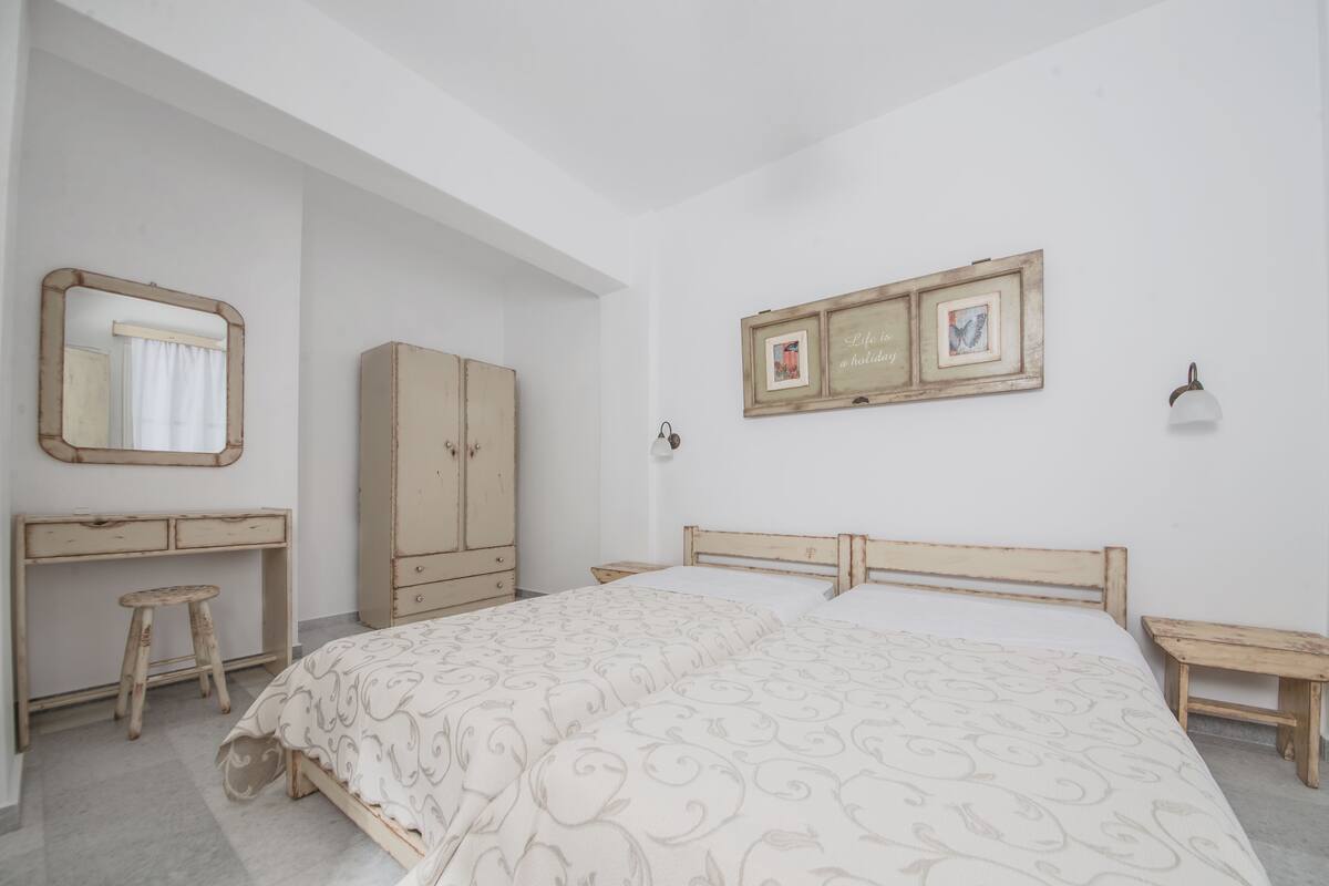 A bedroom with a bed, a wooden wardrobe, a desk with a chair, a mirror, and a wall-mounted shelf with pictures.
