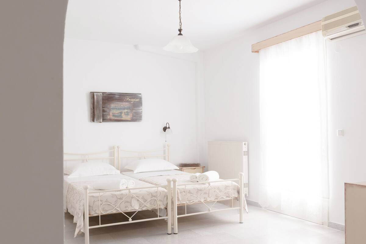 A white, two-bed room with a wooden headboard, a bedside table, a wall-mounted lamp, and a large window with curtains.