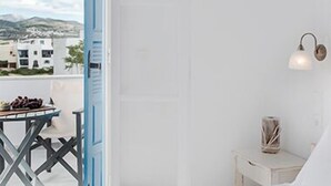 Una camera da letto con un letto, un tavolo con frutta, una lampada a parete e vista su edifici e montagne attraverso una porta aperta.