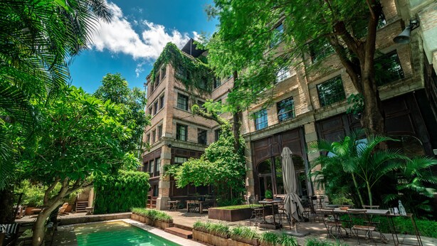 Zona de piscina con mobiliario exterior, una piscina y un edificio de paredes de ladrillo y ventanas amplias.
