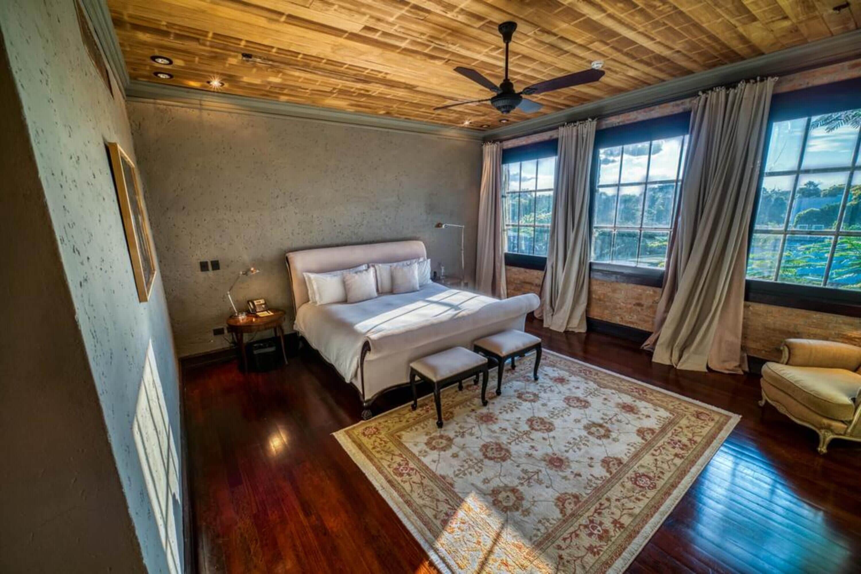 Ein Schlafzimmer mit einem großen Bett, einem Deckenventilator, einem Nachttisch, einem Sessel und Blick ins Grüne durch das Fenster.