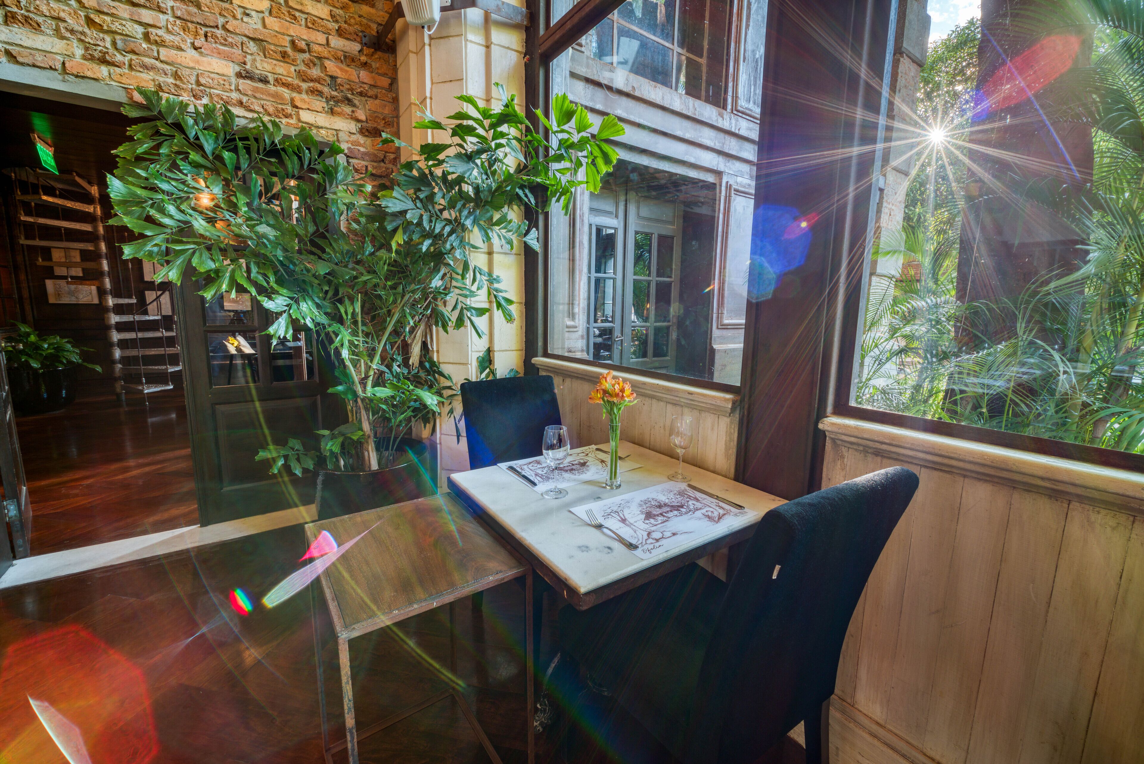 Un comedor con una mesa dispuesta para dos, una planta en maceta y una ventana con vista a follaje.