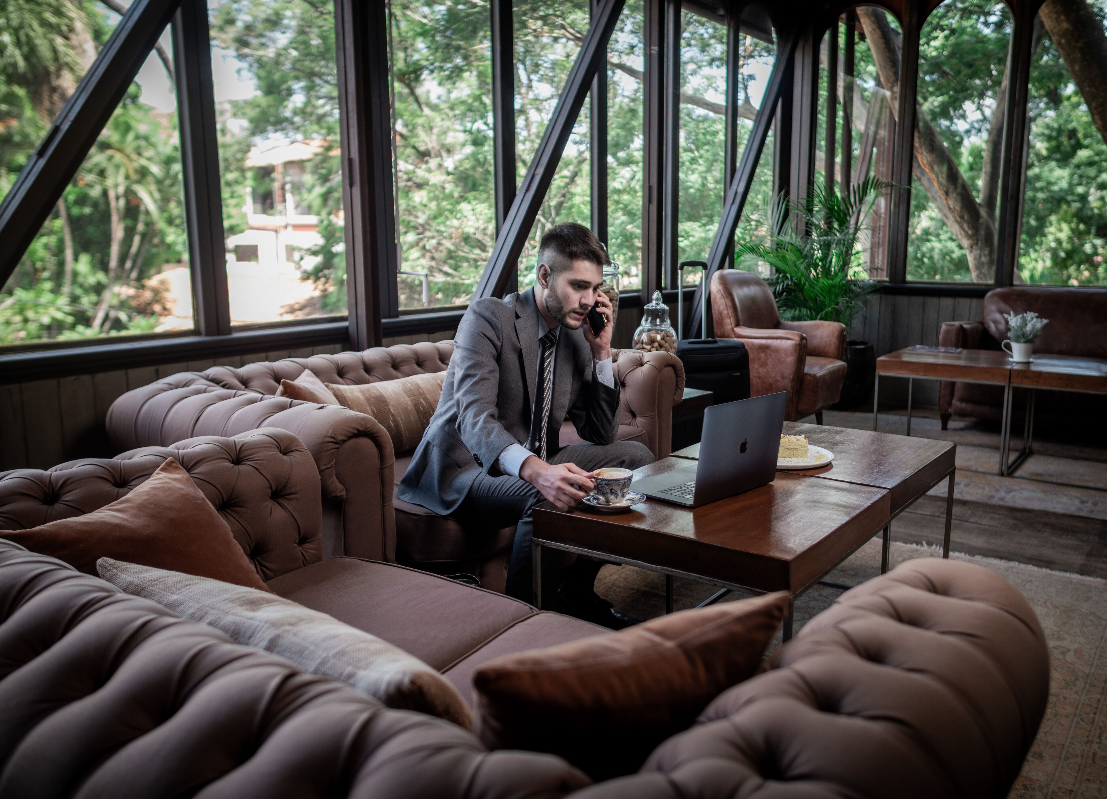 A man in a suit sitting on a tufted leather sofa, using a laptop at a wooden table with a cup of coffee and a plate of food.