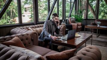 A man in a suit sitting on a tufted leather sofa, using a laptop at a wooden table with a cup of coffee and a plate of food.