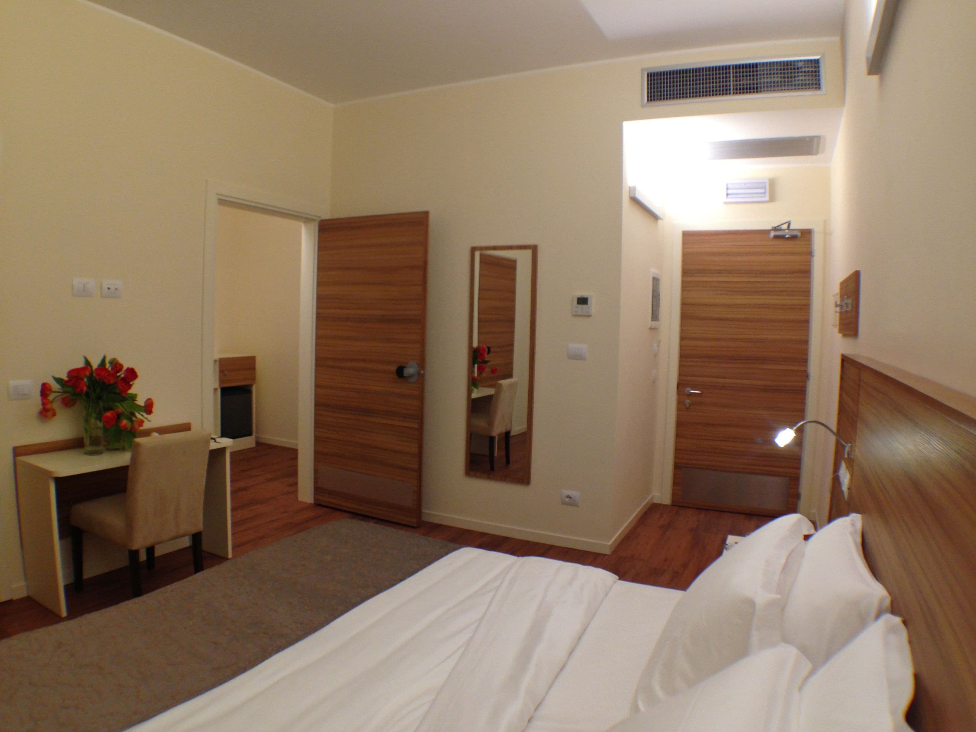 A hotel room with a bed, a desk with a chair, a mirror, and a bathroom door.