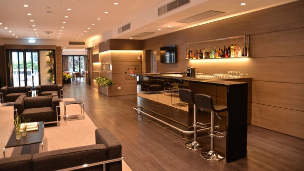 A modern bar area with a long counter, multiple stools, and a variety of bottles. There are sofas and a small table with a vase in the foreground. - Hotel Forum (Baranzate)