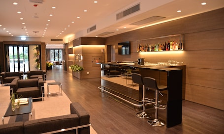A modern bar area with a long counter, multiple stools, and a variety of bottles. There are sofas and a small table with a vase in the foreground.