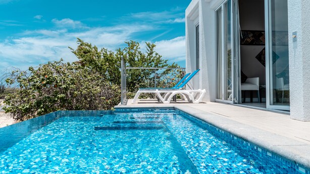 Ein Schwimmbecken mit klarem, blauen Wasser, umgeben von einem weißen Gebäude mit großen Glasfenstern und einem weißen Liegestuhl am Pool.