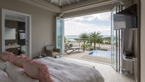 Ein Schlafzimmer mit einem großen Bett, einem Schreibtisch und einem Fernseher. Es besitzt einen Balkon mit Pool und Blick auf den Strand.