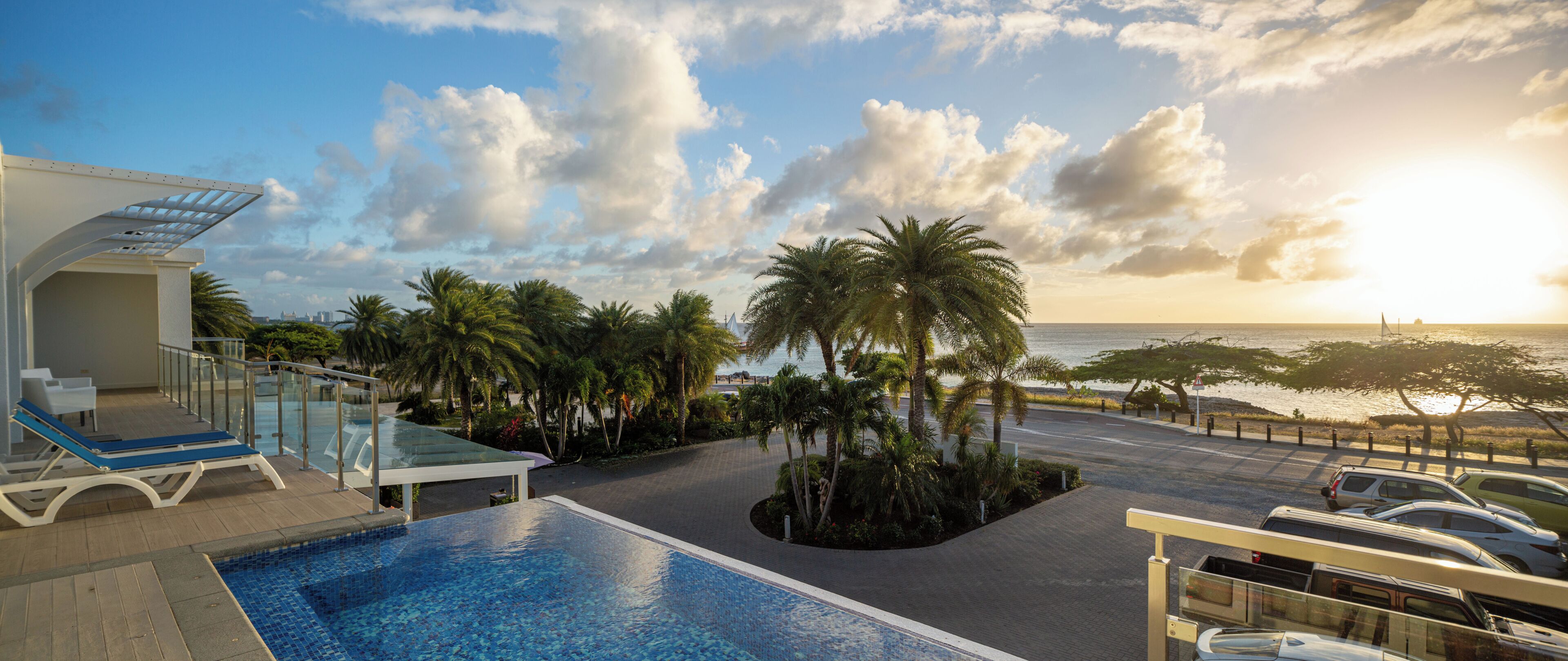 Zona de piscina con sillones, palmeras y vista al océano al atardecer.
