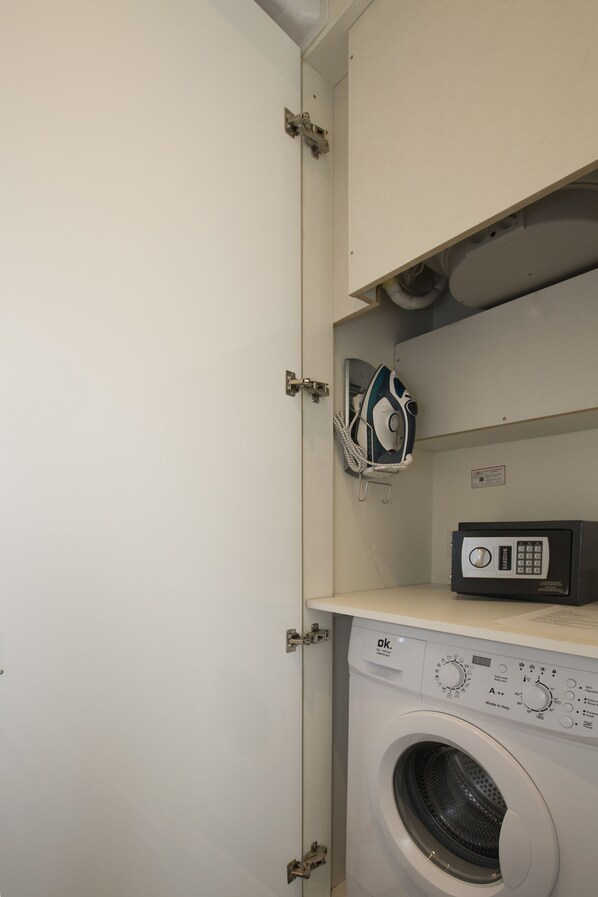 A compact laundry room with a front-loading washing machine, a wall-mounted ironing board, and a small heater on the shelf.
