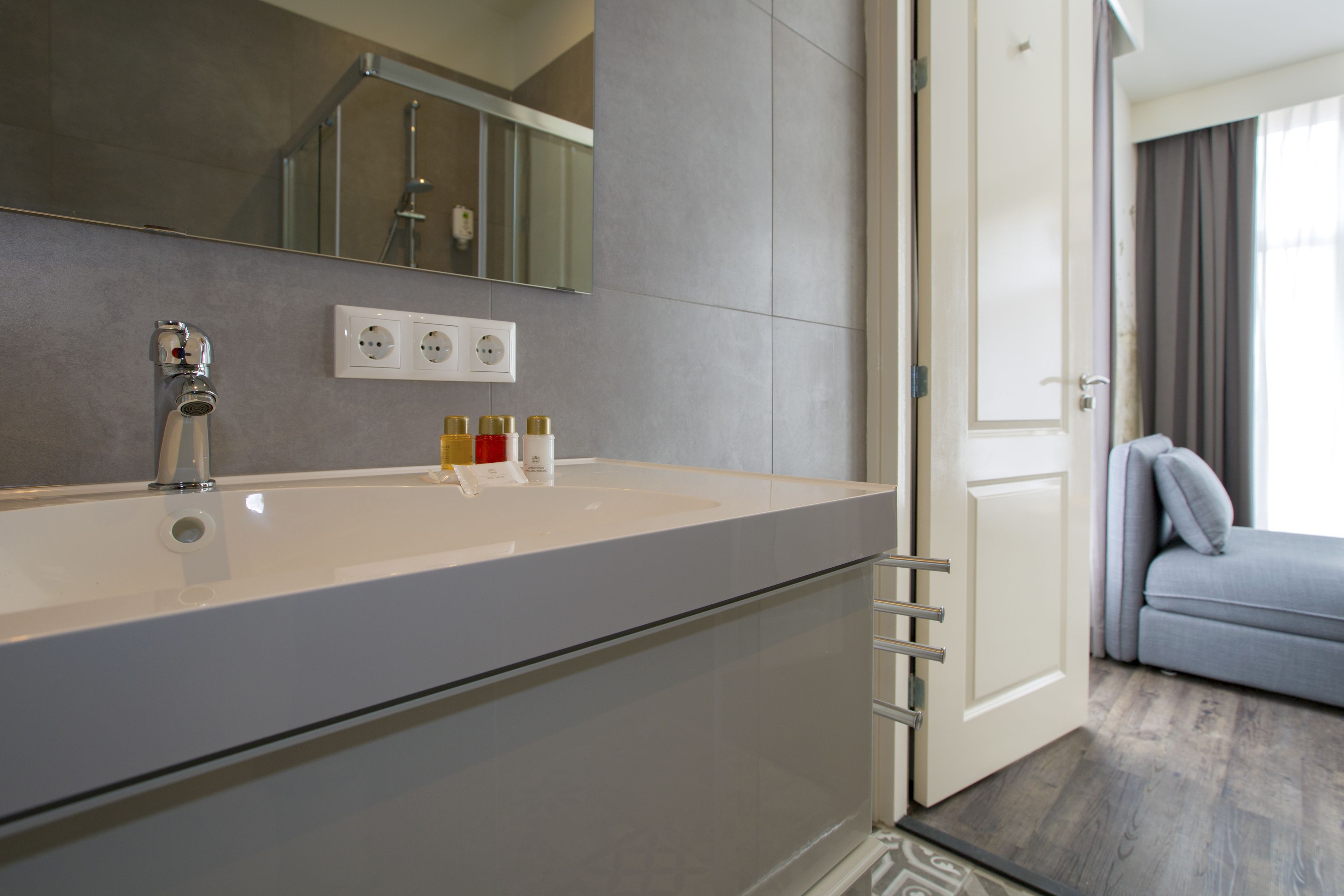Ein modernes Badezimmer mit einem großen Waschbecken, einem Spiegel und einer Dusche. Auf dem Waschbecken stehen Pflegeprodukte und eine Tür führt in einen Wohnbereich.