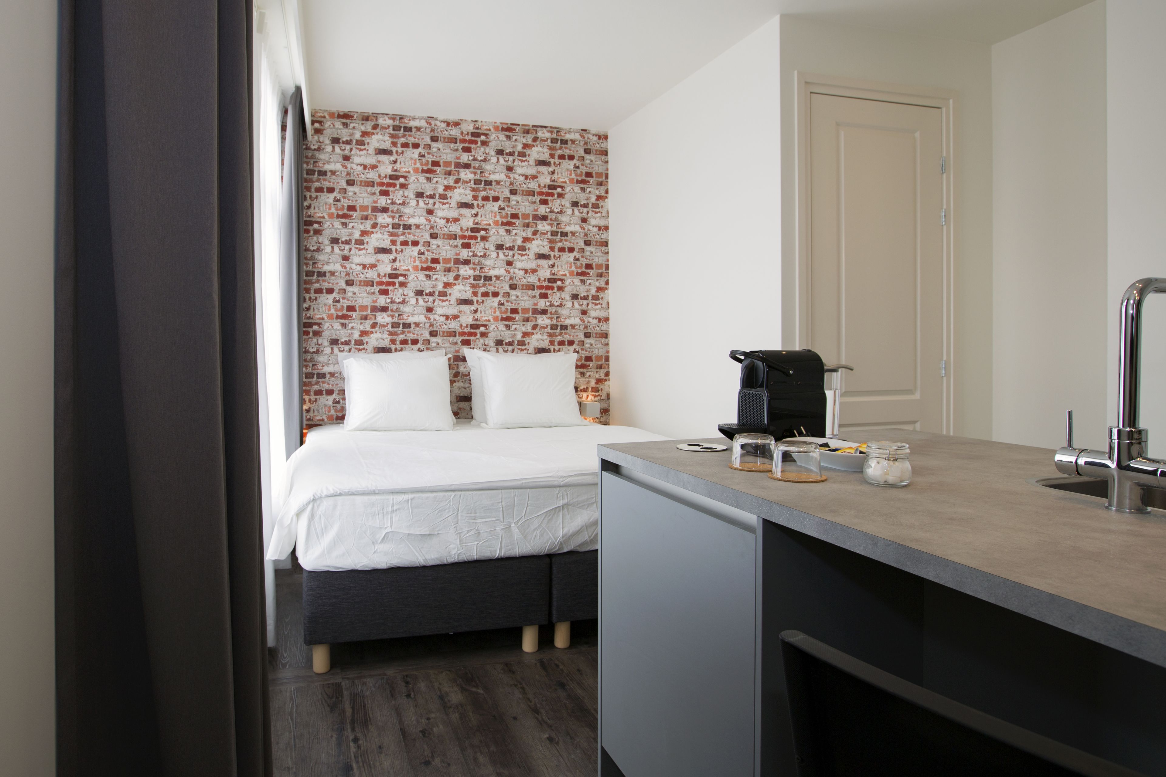 A compact hotel room with a bed, a small kitchenette, and a brick-patterned wall.