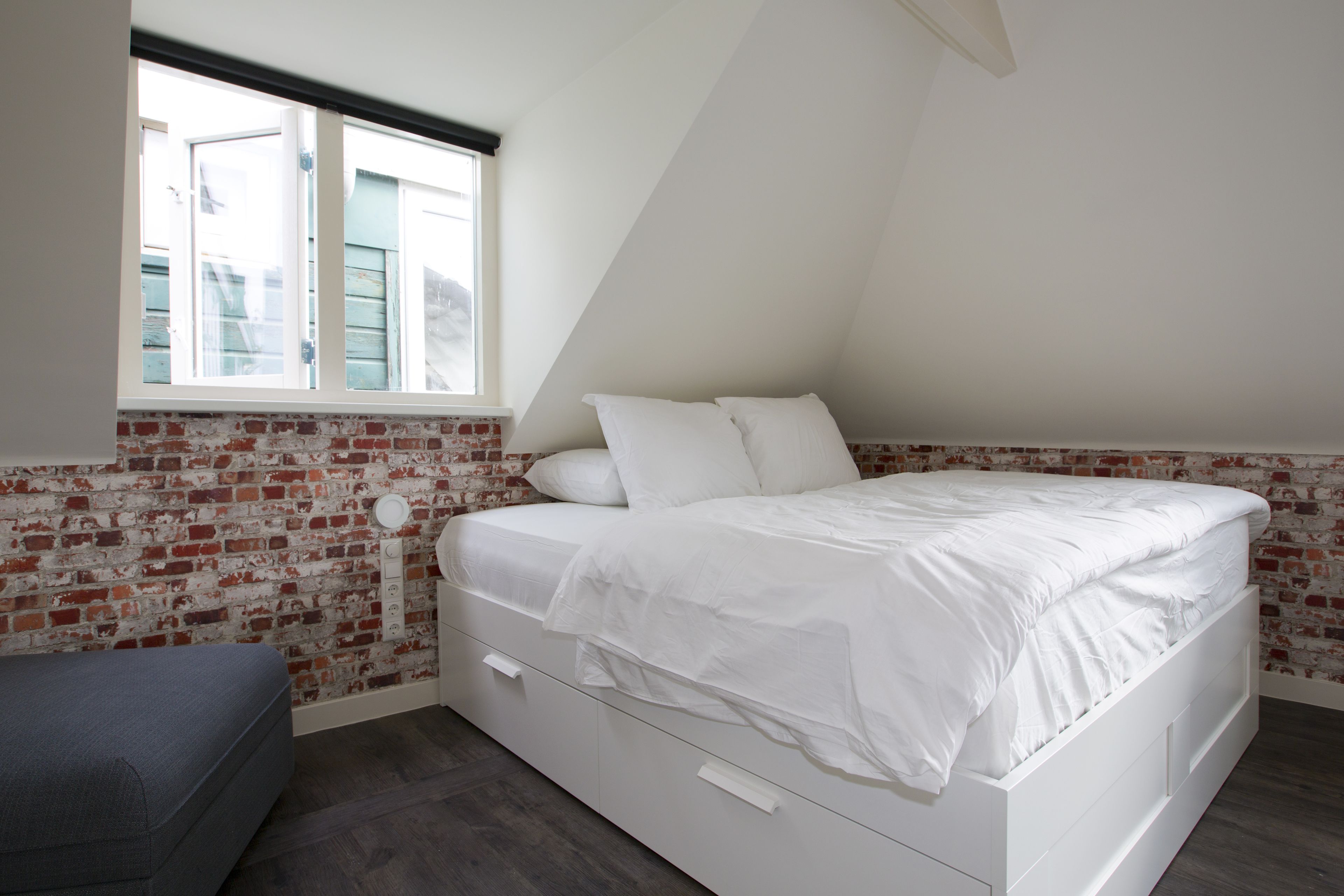 Quarto com cama branca, um banco cinza, parede com padrão de tijolos e janela com persianas.