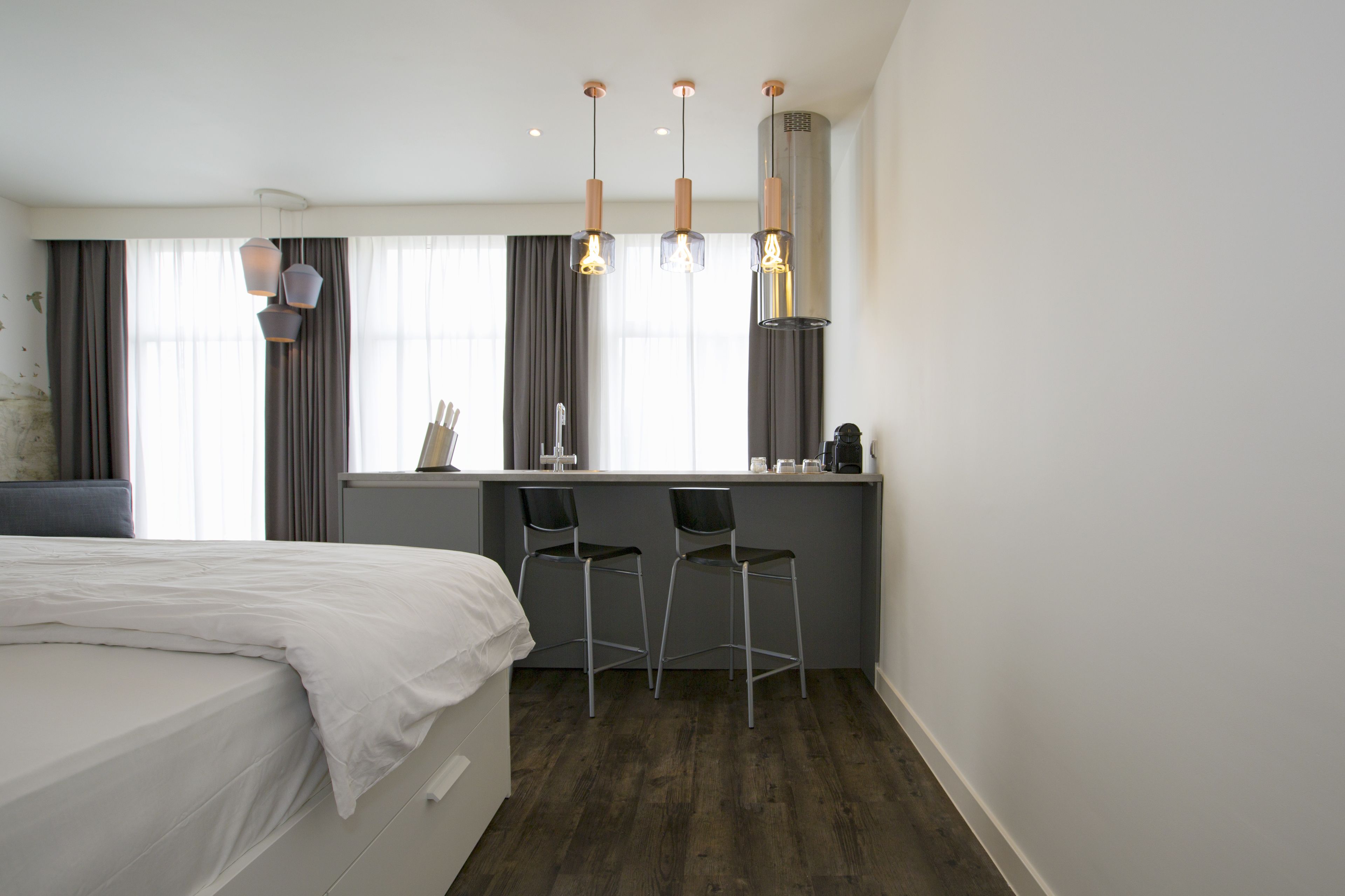 Een moderne hotelkamer met een bed, een klein kitchenette, twee barkrukken en een groot raam met gordijnen.